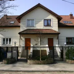 Jasny bliźniak z brązowym dachem i dwoma wejściami, otoczony czarnym, kutym ogrodzeniem.