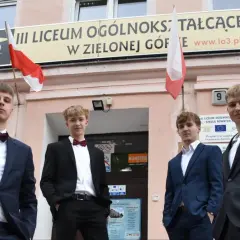 Czterech młodych mężczyzn w garniturach przed budynkiem III Liceum Ogólnokształcącego w Zielonej Górze.
