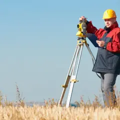 Geodeta w żółtym kasku i kurtce pracuje z niwelatorem. Zastanawiasz się, ile kosztuje geodeta przy budowie domu?