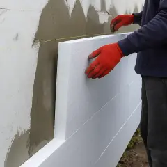 Ręce w czerwonych rękawicach przyklejają styropian do ściany. Sprawdzamy cenę robocizny za klejenie styropianu.