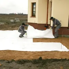 Dwóch robotników rozkłada białą włókninę na placu budowy obok domu.