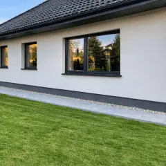 Nowy dom z zadbanym trawnikiem i ścieżką z płyt. Opaska wokół domu z kamyczków chroni elewację.