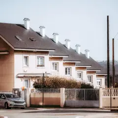 Rządowy dom szeregowy z białymi kominami i ogrodzeniem. Obok stoi srebrny samochód.