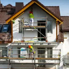 Pracownicy na rusztowaniach prowadzą termomodernizację budynku, ocieplając jego fasadę.
