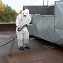 Pracownik w białym kombinezonie natryskuje papę na zimno na dachu.
