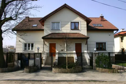 Jasny bliźniak z brązowym dachem i dwoma wejściami, otoczony czarnym, kutym ogrodzeniem.
