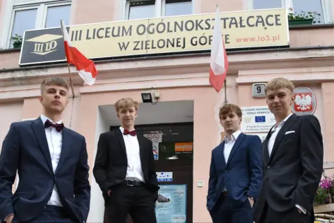 Czterech młodych mężczyzn w garniturach przed budynkiem III Liceum Ogólnokształcącego w Zielonej Górze.