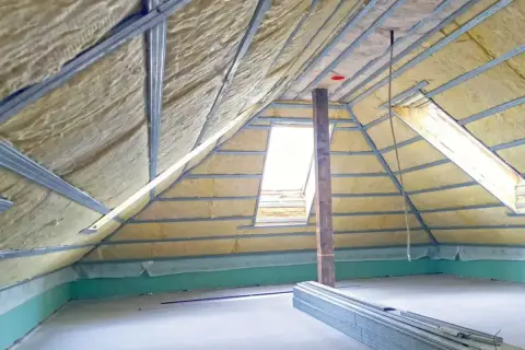 Izolowane poddasze nieużytkowe z widocznymi elementami konstrukcyjnymi i oknem dachowym. Widać materiały budowlane.