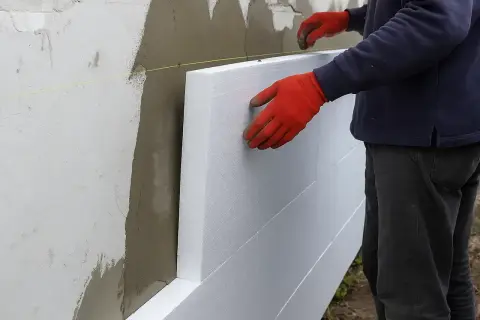 Ręce w czerwonych rękawicach przyklejają styropian do ściany. Sprawdzamy cenę robocizny za klejenie styropianu.