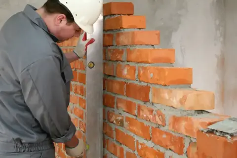 Murarz w białym kasku sprawdza pion muru, używając poziomicy. Widać cegły o standardowych wymiarach, układane na zaprawie.