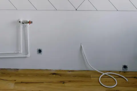 Białe rury wodne i zawór na ścianie, gniazdka elektryczne i biały kabel zwinięty na drewnianej podłodze. Instalacja gazowa w domu w trakcie prac.