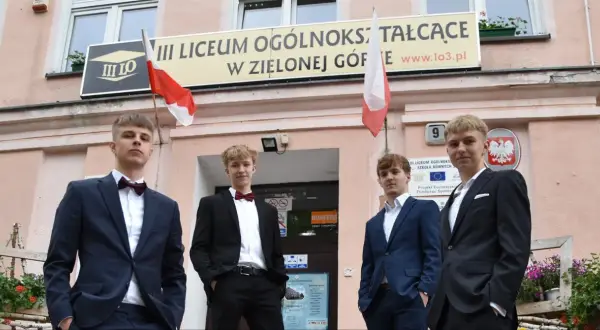 Czterech młodych mężczyzn w garniturach przed budynkiem III Liceum Ogólnokształcącego w Zielonej Górze.