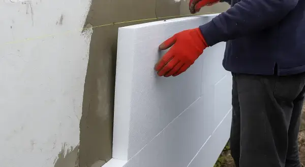 Ręce w czerwonych rękawicach przyklejają styropian do ściany. Sprawdzamy cenę robocizny za klejenie styropianu.