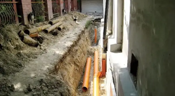 Trwa budowa drenażu wokół domu. W wykopie leżą nowe pomarańczowe rury i stare, zniszczone rury.