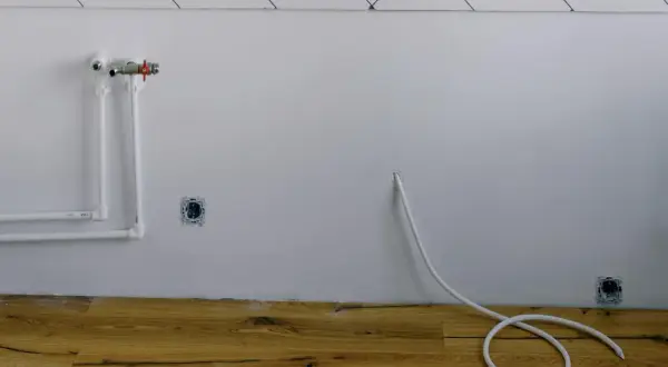 Białe rury wodne i zawór na ścianie, gniazdka elektryczne i biały kabel zwinięty na drewnianej podłodze. Instalacja gazowa w domu w trakcie prac.