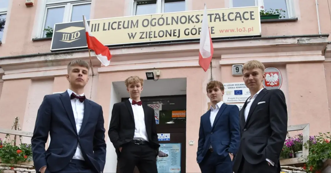 Czterech młodych mężczyzn w garniturach przed budynkiem III Liceum Ogólnokształcącego w Zielonej Górze.