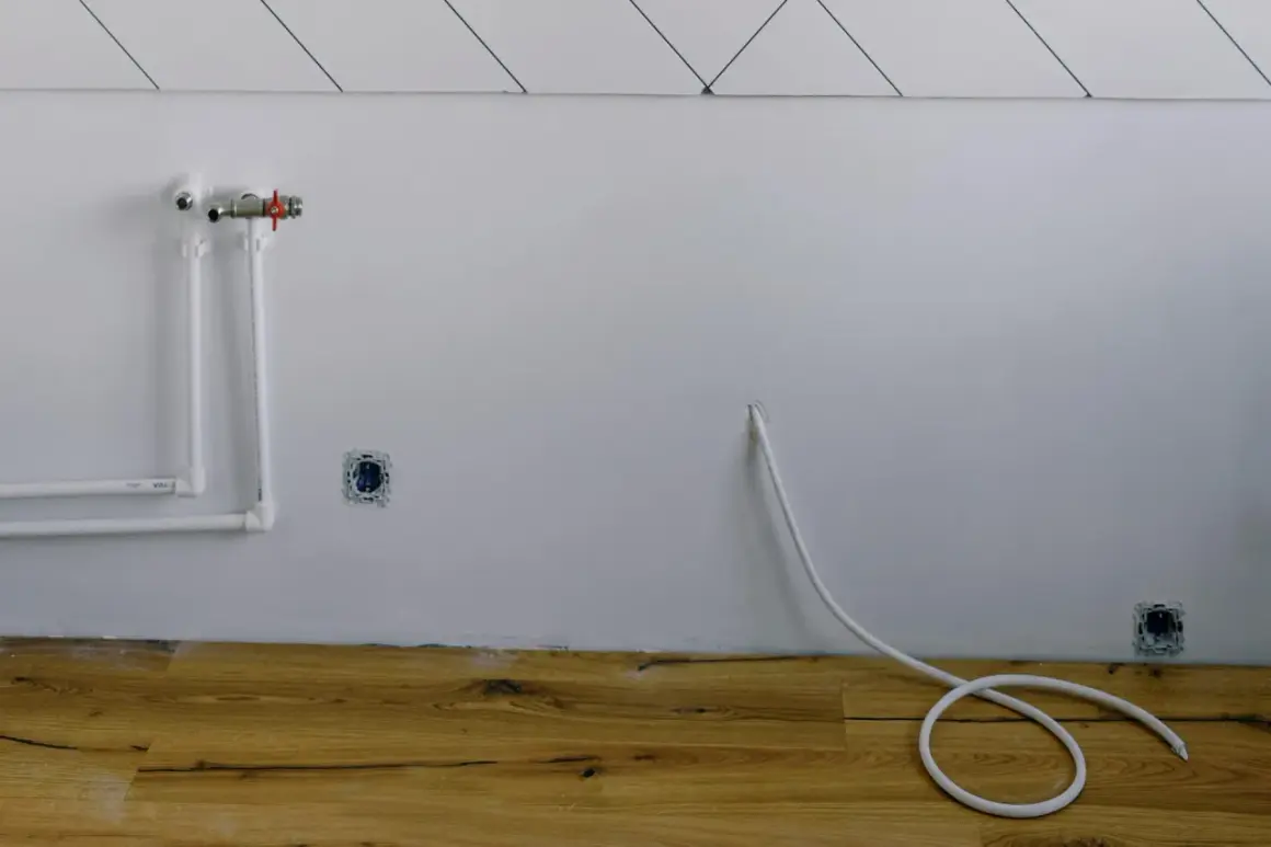 Białe rury wodne i zawór na ścianie, gniazdka elektryczne i biały kabel zwinięty na drewnianej podłodze. Instalacja gazowa w domu w trakcie prac.