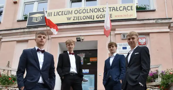 Czterech młodych mężczyzn w garniturach przed budynkiem III Liceum Ogólnokształcącego w Zielonej Górze.